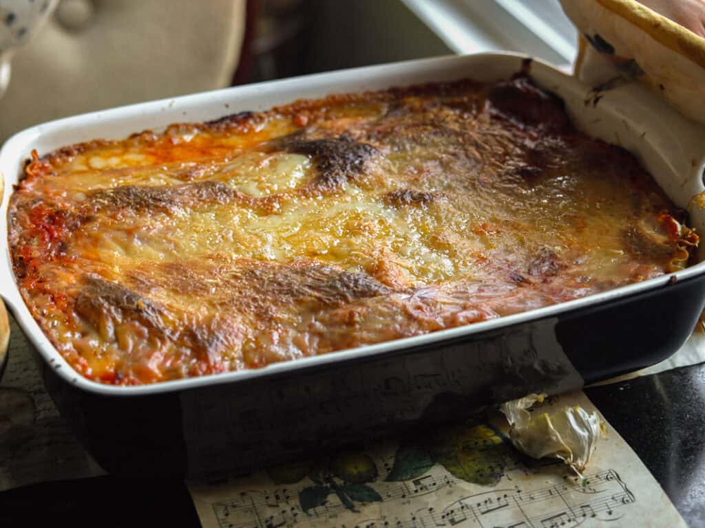 A baked vegan lasagna in a black and white rectangular dish, featuring a golden-brown and slightly crispy cheese topping. It is placed on a table with a printed sheet and a partially visible bread basket nearby.